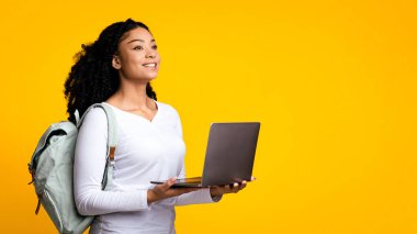 Online Eğitim Konsepti. Sırt çantalı ve dizüstü bilgisayarlı genç siyahi bir kızın portresi sarı arka planda izole edilmiş, Afro-Amerikan bir kadının bakışları uzaklara bakıyor ve gülümsüyor,