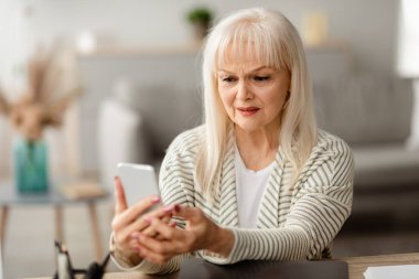 Mutsuz, orta yaşlı bir kadın telefon ekranına bakıyor, oturma odasında oturuyor, sosyal ağda kötü haber okuyan sinirli yaşlı bir kadın, cihazla ilgili bir sorun var.