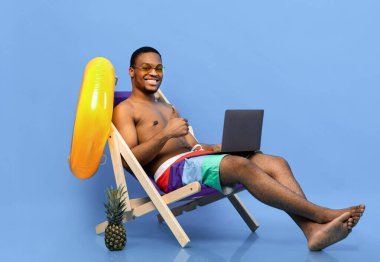 Mayo giyen neşeli siyah adam dizüstü bilgisayar kullanıyor, plaj tatili sırasında internette geziniyor, şezlongda oturuyor, mavi stüdyo arka planında baş parmak hareketlerini gösteriyor. Yaz tatili kavramı