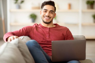 Evdeki dizüstü bilgisayarla koltuğa yaslanmış gülümseyen Arap adam, kopyalama alanı. Yakışıklı, orta doğulu genç bir adam hafta sonu eğleniyor, defter kullanıyor, 