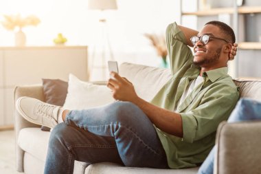 Yeni Mobil Uygulama. Neşeli siyah adam cep telefonu kullanıyor, gözlük takıyor, mesaj atıyor ve evdeki koltukta oturup internete bakıyor. İçerde Kullanıcı Kullanımı Uygulaması. Yan Görünüm
