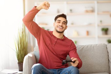 Duygusal ortadoğulu genç adam evde joystick ile video oyunu oynuyor, oturma odasında kanepede oturuyor, hafta sonları eğleniyor, mekanı kopyalıyor. Ev eğlencesi, eğlence, ev hayatı.