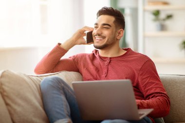 Coronavirüs salgını sırasında evde çalışan, dizüstü bilgisayar kullanan ve cep telefonuyla konuşan, oturma odasındaki kanepede oturup fotokopi çeken, orta doğulu sakallı iş adamı.