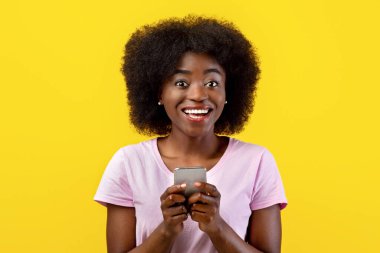 Yeni havalı uygulama. Akıllı telefonunu kullanan mutlu zenci kadının portresi, internete göz atmak, kameraya mesaj atmak ve gülümsemek, sarı stüdyo arka planının başında durmak,