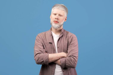 Genç albino sarışın adam kollarını kavuşturup poz veriyor, günlük kıyafetler giyiyor ve turkuaz stüdyo arka planından kameraya bakıyor. Yüzünde ciddi bir ifade olan adam. Albinizm kavramı