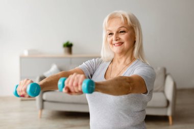 Ev Fizyoterapi Konsepti. Gülümseyen Yaşlı Kadın 'ın portresi Oturma odasında iki dambılla egzersiz yapıyor, spor yapıyor. Boş Kopyalama Alanı, Bulanık Arkaplan, Seçici Odak