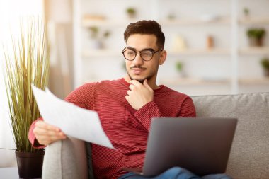 Konsantre olmuş Arap işadamı ya da serbest çalışan, kanepede oturan ve oturma odasında dizüstü bilgisayar kullanan, gazete okuyan, çenesine dokunan, gözlük takan, fotokopi çeken