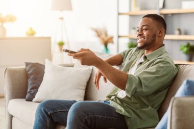 Mutlu Afro-Amerikan Adam TV izlerken uzaktan kumandalı televizyon kanallarını değiştirip evdeki koltukta otururken yan görüntüsü. Haftasonu Eğlencesi, TV Programı Konsepti