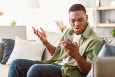 Smartphone 'da sorun var. Cep telefonu kullanan hoşnutsuz Afro-Amerikalı adamın evdeki koltukta oturan cep telefonu uygulaması sorunu var. Memnun Olmayan Telefon Kullanıcı Okuma Negatif Haberler Çevrimiçi