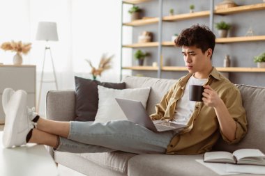 Ev İşyeri. Genç ciddi Asyalı adam oturma odasında çalışıyor, kanepede dizüstü bilgisayarla oturuyor, kahve içiyor. Bilgisayar kullanan, iş haberleri okuyan, kanepede dinlenen profesyonel bir adam.