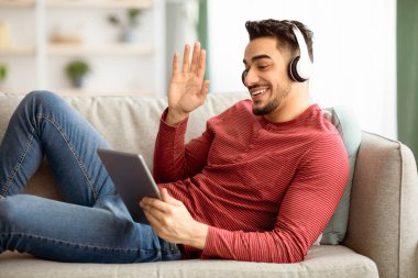 Neşeli genç Müslüman adam arkadaşlarıyla ya da sevgilileriyle video konferansı yapıyor, dijital tablet ve kablosuz kulaklık kullanıyor, tablet ekrana bakıyor, gülümsüyor ve el sallıyor, ev içini kopyalıyor.