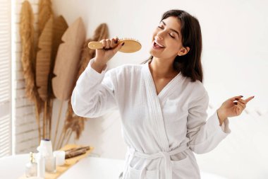 Saç Bakımı. Neşeli esmer kadın saç fırçasıyla şarkı söylüyor modern banyoda eğleniyor. Saç bakımı kozmetik reklamı. Güzellik Ayinleri Konsepti