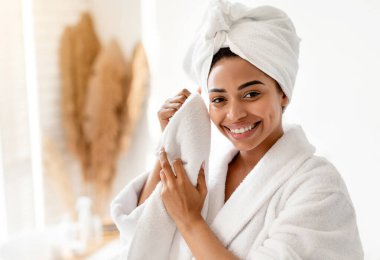 Yüzü havluyla kurumuş genç siyah bir kadın cilt bakımı yapıyor modern banyoda kameraya gülümsüyor. Şımartma, Wellnes ve Spa. Dişi, Güzelliğin Zevkini Çıkartıyor