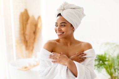 Çekici Afro-Amerikan kadın pozu çıplak omuzlu, beyaz bornoz giyiyor ve evde banyoda duran mükemmel vücuda özen gösteriyor. Sağlık ve Spa Konsepti