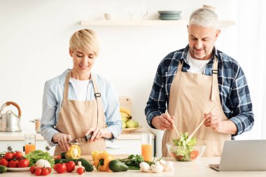 Çift sağlıklı vejetaryen salatası hazırlıyor, modern rahat mutfağın tadını çıkarıyor, birlikte yemek pişiriyor, evde keyif çatıyor. Masada sebzeli önlüklü, gülümseyen yetişkin kadın ve adam.
