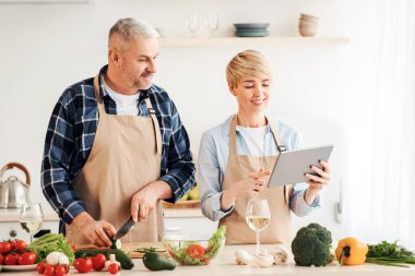 Evde modern teknolojilerle sağlıklı yemekler hazırlayın. Mutlu yetişkin erkek sebze keser, kadın mutfak içinde tablete bakar masada parlak sebzeler ve şarap kadehleri, boş alan