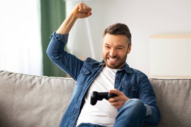 Duygusal orta yaşlı sakallı bir adam evde joystick ile video oyunu oynuyor, oturma odasında kanepede oturuyor, hafta sonları eğleniyor, mekanı kopyalıyor. Ev eğlencesi, eğlence, ev hayatı.