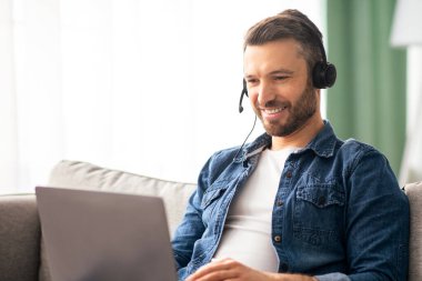 Positif sakallı adam evden video konferansı yapıyor, kulaklık ve laptop kullanıyor, konuşuyor ve gülümsüyor. Keyifli orta yaşlı bir adam oturma odasındaki koltukta dinleniyor, görüntülü sohbet ediyor, boşluğu kopyalıyor.