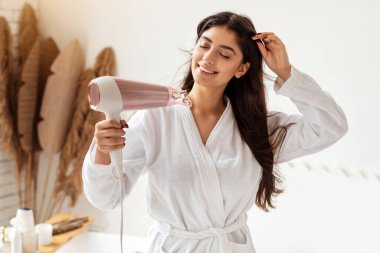 Güzel esmer, kuruyan ve saç kurutma makinesi pozu veren bir kadın. Gözleri kapalı, banyoda dikiliyor. Sabah duşundan sonra saç kurutma makinesi kullanan kadın. Kadın Saç Bakımı Konsepti