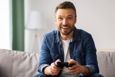 Duygusal orta yaşlı sakallı bir adam evde joystick ile video oyunu oynuyor, oturma odasında kanepede oturuyor, hafta sonları eğleniyor, fotokopi çekiyor. Ev eğlencesi, eğlence, ev hayatı.