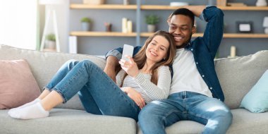 Neşeli çok kültürlü eşler akıllı telefonlarıyla koltukta dinleniyorlar, mutlu ırklar arası aşıklar evdeki oturma odasında rahat bir kanepede dinlenirken cep telefonu kullanıyorlar, boşluğu kopyalıyorlar.