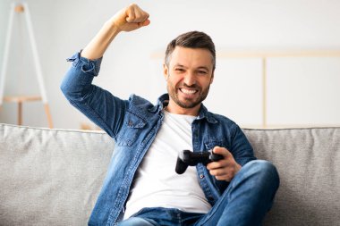 Duygusal orta yaşlı sakallı bir adam evde joystick ile video oyunu oynuyor, oturma odasında kanepede oturuyor, hafta sonları eğleniyor, mekanı kopyalıyor. Ev eğlencesi, eğlence, ev hayatı.