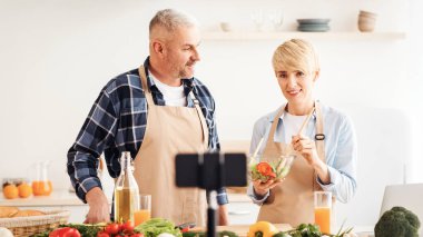 Çift birlikte yemek pişiriyor, yemek tarifi veriyor, video bloğu ve sosyal medya videolarını akıllı telefon kamerası ve tripodla kaydediyor. Mutlu bir eş ve koca mutfak içinde yemek hazırlar, boş bir alan, bulanık.