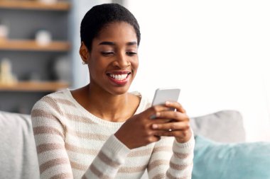 Mobil İletişim. Evde akıllı telefondan mesajlaşan genç siyahi kadın, oturma odasında dinlenirken arkadaşlarıyla cep telefonunda mesajlaşan neşeli Afrikalı Amerikalı kadın,