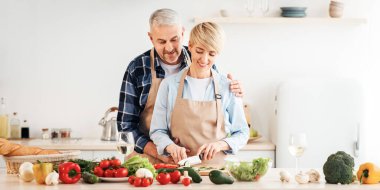 Sağlıklı bakım, yararlı yaşam tarzı, düzgün yemek, vejetaryen ve ev yapımı yemek. Gülümseyen olgun bir kadın yemek pişiriyor, erkek mutfakta kadına sarılıyor. Parlak sebzelerle, masada bir kadeh şarapla, boş bir alanda.
