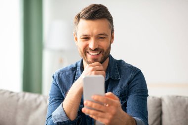 Sevgilisiyle görüntülü sohbetten hoşlanan mutlu orta yaşlı adam cep telefonu ekranına bakıyor ve gülümsüyor, fotokopi çekiyor. Oturma odasında kanepede otururken elinde akıllı telefon tutan neşeli sakallı adam.
