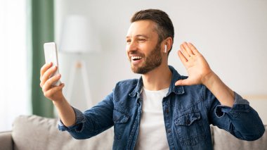 Olumlu orta yaşlı bir adam COVID-19 salgını sırasında evde otururken arkadaşlarıyla ya da sevgilileriyle online parti veriyor, koltukta oturuyor ve kablosuz kulaklık ve cep telefonu kullanıyor.