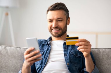 Kredi kartı ve cep telefonu olan neşeli orta yaşlı bir adam evdeki koltukta oturuyor, yakın plan, kopyalama alanı. Mutlu sakallı adam internetten alışveriş yapıyor, akıllı telefon kullanıyor ve kart tutuyor, e-ticaret konseptini kullanıyor.
