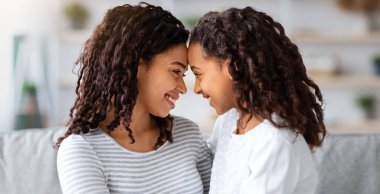 Güzel kıvırcık saçlı, tatlı siyah anne ve kızı oturma odasında kanepede oturmuş, birbirlerine sarılıyorlar. Mutlu Afro-Amerikan anne ve çocuk kucaklaşıp birbirine alnıyla dokunuyor.