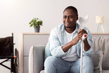 İyileşme, rehabilitasyon, terapi ve olumlu insani duyguların ifadesi. Gülümseyen orta yaşlı Afrikalı Amerikalı koltuk değnekleriyle engelli gri duvarlı minimalist oturma odasında oturuyor.