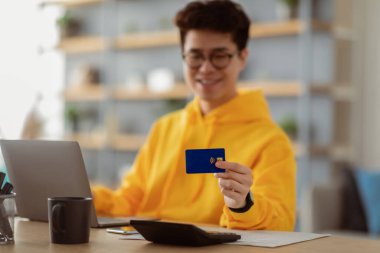 Finansal işlem yapıyor. Gözlüklü gülümseyen genç Asyalı adam bankamatik kartı gösteriyor, evinde dizüstü bilgisayarla masada oturuyor, seçici odaklanma, bulanık bir arka plan.