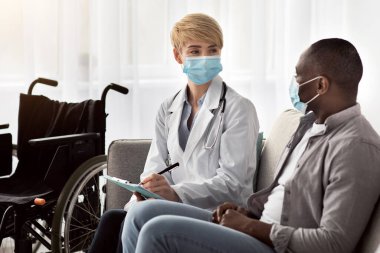 COVID-19 sırasında evdeki tıbbi muayene, engelli kişiyi ziyaret. Kadın doktor ve yetişkin Afro-Amerikalı koruyucu maskeli adam konuşuyor, koltukta ya da klinikte tekerlekli sandalyeli oturuyor.