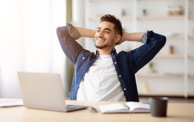 Mutlu genç Arap adam işini seviyor, çalışma masasında oturuyor, arkasına yaslanıyor, fotokopi odasına bakıyor ve gülümsüyor, ofis içi. Milenyum için iş fırsatları, kariyer kavramı