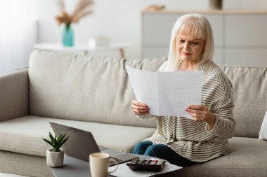 Evrak işi kavramı. Evde çalışan, elinde kağıt ya da finansal belge tutan, oturma odasında dizüstü bilgisayarla oturan, boş alan olan olgun bir kadının portresi.