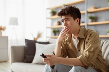 Evde Sıkıcı Film. Uzaktan kumandayla televizyon izleyen yorgun Asyalı bir adamın portresi, esniyor, ağzı elle kapanıyor, oturma odasında kanepede oturuyor. Covid19 Karantina, Tecrit, Tecrit