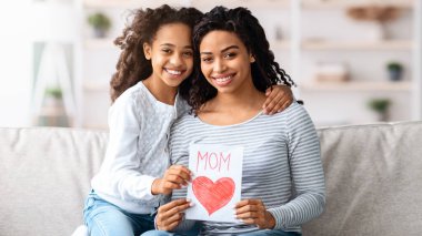 Annesine sarılan mutlu siyah kız, annesini Anneler Günü 'nde karşılayan sevgili kızı, el sanatları hediyesini tutan genç kadın, kameraya gülümseyen, ev içi, Anneler Günü kutlaması.