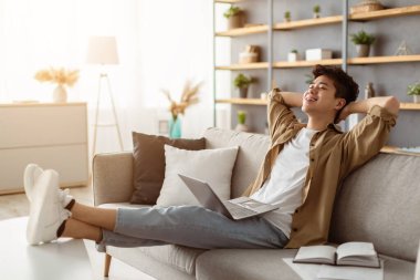 Ara veriyoruz. Gülümseyen Asyalı genç adam oturma odasında rahat bir kanepede dizüstü bilgisayar kullanarak rahatlıyor, mutlu milenyum erkeği evdeki rahat kanepede dinleniyor, arkasına yaslanıyor, hafta sonunun keyfini çıkarıyor ya da serbest çalışıyor.