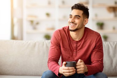 Kanepede oturup çay ya da kahve içen neşeli bir Arap. Orta Doğulu genç adam elinde kupayla evde vakit geçirip gülümsüyor ve fotokopi odasına bakıyor.