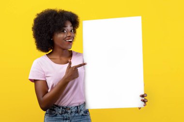 Mesaj için yer. Heyecanlı Afro-Amerikalı bayan sarı arka planda tasarladığınız yer olan boş pankartı işaret ediyor. Pozitif siyah kadın boş poster gösteriyor, yaz reklamı için maket yapıyor.
