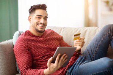 Kredi kartı ve dijital tabletle evdeki kanepede dinlenirken internetten alışveriş yapan mutlu genç bir Arap. Gülümseyen orta doğulu adam satın alıyor, ped, e-ticaret, yan görüş kullanıyor.