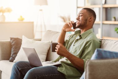 Cep telefonuyla konuşan, dizüstü bilgisayar kullanan ve evdeki koltukta oturup kahve içen mutlu siyahi adam. Modern Mobil İletişim, Telefon Çağrısı. Her zaman temastayım. Yan Görünüm