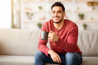 Kanepede oturup çay ya da kahve içen neşeli bir Arap. Orta Doğulu genç adam elinde kupayla elinde fotokopi odasına bakarak evde vakit geçiriyor.
