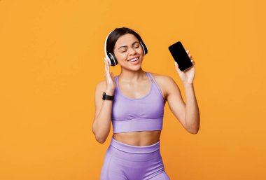 Egzersiz sırasında favori şarkının tadını çıkarıyorum. Kablosuz kulaklık takan, elinde boş ekranlı akıllı telefon tutan, sarı arka planda duran siyahi bir kadın.