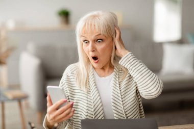 Evde akıllı telefondan şok edici haberler okuyan endişeli yaşlı bayanın portresi, ekrana bakıyor ve şok içinde başını tutuyor, boş alan. Siber Dolandırıcılık, Mobil Dolandırıcılık