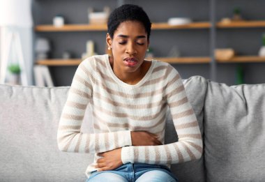 Mide Ache. Evde şiddetli karın ağrısı çeken siyah bir kadın koltukta otururken karnına dokunan Afrikalı genç bir kadın regl sancısı çekiyor ya da sindirim sorunları yaşıyor.