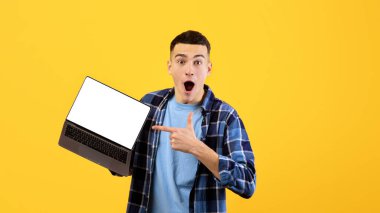 Portakal rengi stüdyo arka planında web sitesi tasarımı için yeri olan boş dizüstü bilgisayarı işaret eden genç adam şok oldu. Heyecanlı bir milenyum adamı reklamınız için taşınabilir bilgisayarını gösteriyor.